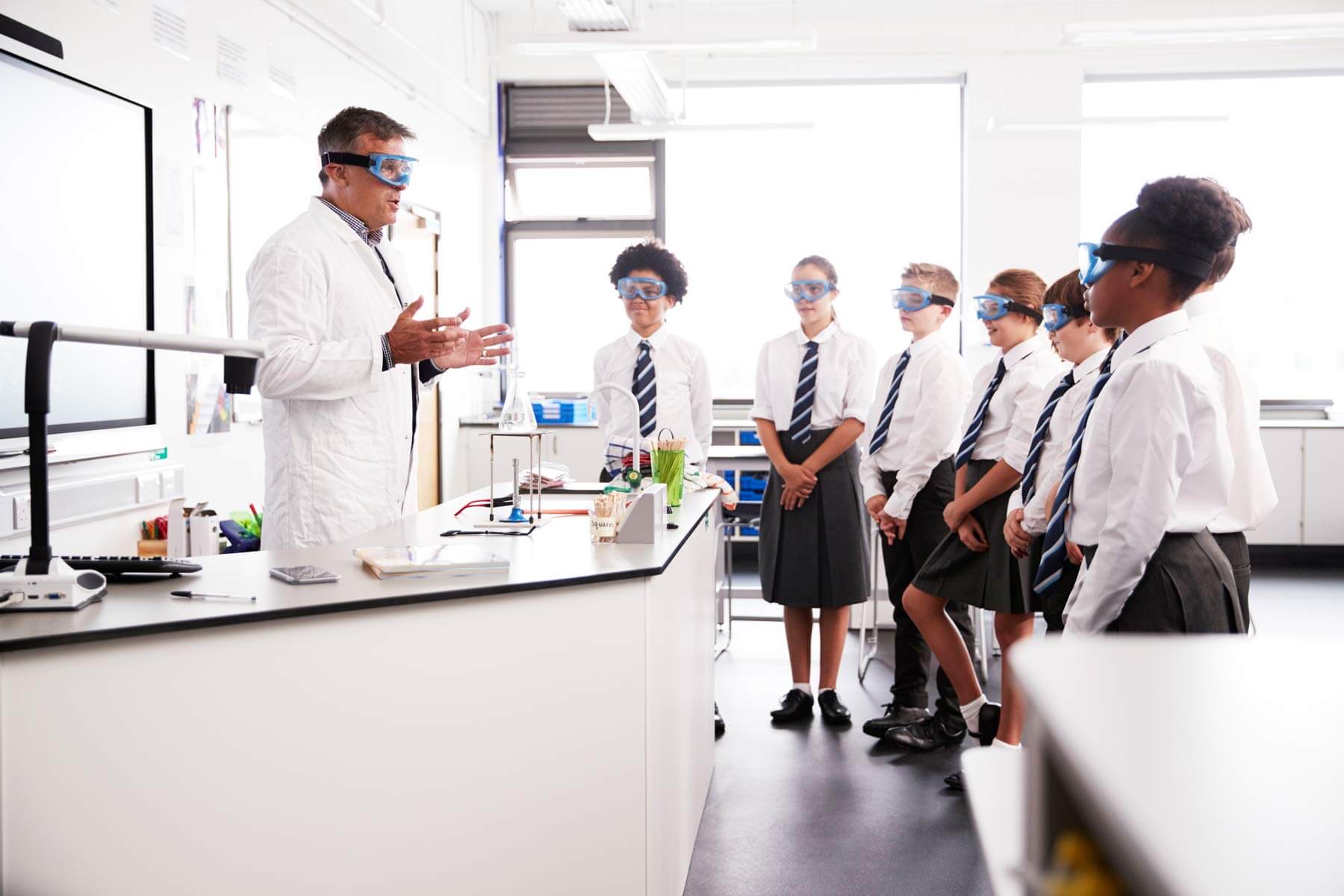 Teacher in lab coat talks to students wearing goggles